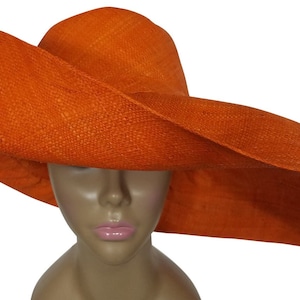 May include: A large, wide-brimmed hat made of woven straw in a bright orange colour. The hat is tilted slightly to the side, and the brim is curved and flowing.