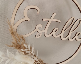 Puede incluir: Un círculo de madera con el nombre "Estelle" escrito en cursiva. El círculo está decorado con flores secas blancas y marrones.