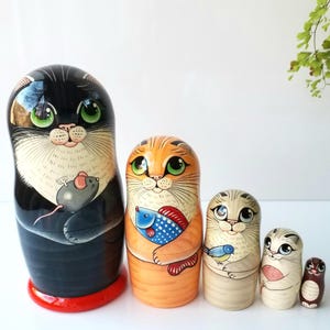 May include: A set of five wooden nesting dolls, each painted with a cat design. The largest doll is black with a mouse, the next is orange with a fish, followed by a gray cat with a bird, a smaller gray cat, and a small brown doll.
