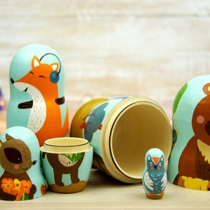May include: A set of six wooden nesting dolls, or matryoshka dolls, painted with whimsical woodland animals. The largest doll is a brown bear, followed by a fox, a raccoon, a deer, a hedgehog, and an owl.