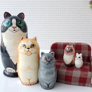 May include: A collection of hand-painted wooden nesting dolls, each shaped and decorated to resemble a different cat breed. The dolls range in size and feature various colors, including black, white, orange, gray, and brown. One doll sits on a miniature plaid couch.