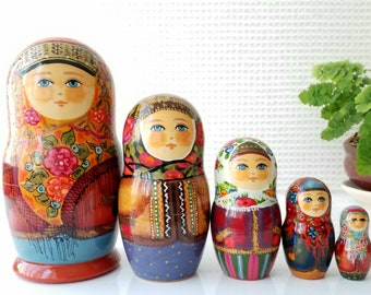 May include: A set of five traditional Russian nesting dolls, or matryoshka dolls. The dolls are painted in bright colors with intricate designs and floral patterns. The largest doll is wearing a red and blue dress with a floral scarf, while the smaller dolls are dressed in similar styles.