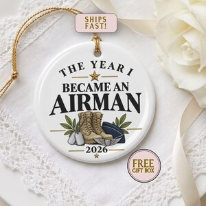 May include: A round white ceramic ornament featuring the text THE YEAR I BECAME AN AIRMAN 2026. The design includes a gold star, tan military boots, silver dog tags, a blue flight cap, and green leaves, suspended by a gold cord.
