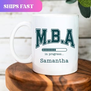 May include: White ceramic mug with a large handle. The mug features the text "M.B.A" in a dark teal collegiate font, with "in progress..." and the name "Samantha" below. A loading bar graphic is also included.