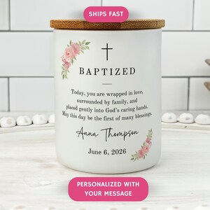 May include: White ceramic candle with a wooden lid, adorned with floral accents and the word "BAPTIZED". The candle includes a cross symbol and personalized text, including a name and date. The design is simple and elegant.
