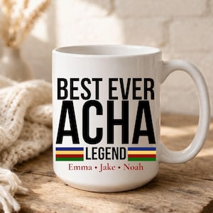May include: White ceramic mug with the text "BEST EVER ACHA LEGEND" in black. Below the text are the names Emma, Jake, and Noah in red. The mug has a handle and is on a wooden surface.