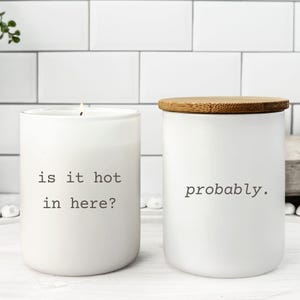 May include: Two white candles with black text. One reads "is it hot in here?" and the other says "probably." The candle on the left is lit. Both candles have a minimalist design and a clean aesthetic.