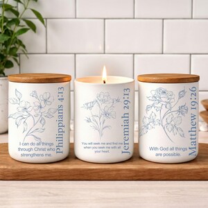 May include: Three white cylindrical candle jars with wooden lids. Each jar features blue floral line art and scripture text: Philippians 4:13, Jeremiah 29:13, and Matthew 19:26. The center candle is lit, displaying a small flame against the white ceramic.