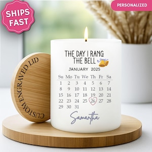 May include: A white candle with a calendar for January 2025, featuring the text "The Day I Rang The Bell" and a heart around the 26th. The candle sits on a wooden stand with a custom engraved lid.
