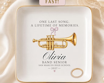 May include: A square white ceramic trinket dish with a gold rim featuring a gold trumpet illustration, a pink bow, and text reading ONE LAST SONG. A LIFETIME OF MEMORIES. Olivia BAND SENIOR SAN MARCOS HIGH SCHOOL 2027 with a small pink heart.