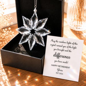 May include: A clear, faceted crystal star ornament suspended from a silver chain inside a black gift box. Beside the box sits a white card with black text: May the rainbow lights of this crystal remind you of the light you brought and the difference you have made! HAPPY RETIREMENT Gloria EST 2024.