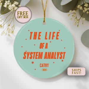 May include: A round, light teal ornament with the text "THE LIFE OF A SYSTEM ANALYST" in orange, along with the name "CATHY" and the year "-2025-". The ornament has a gold string for hanging and is decorated with small orange stars.