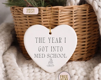 May include: White heart-shaped ornament with the text "THE YEAR I GOT INTO MED SCHOOL" in black. The ornament includes a small graphic and the year "2025". A gold string is attached for hanging. The ornament is in front of a woven basket.