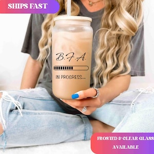May include: A frosted glass tumbler with a bamboo lid and a stainless steel straw. The tumbler is filled with a creamy beverage and features the text "B.F.A. IN PROGRESS..." in black. The tumbler is held by a person.