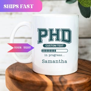 May include: White ceramic mug with a teal "PHD" design, "Custom Text", and a progress bar graphic. The mug also features the name "Samantha" and the text "in progress...". The mug has a large handle and is on a wooden surface.