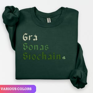 Könnte beinhalten: Ein dunkelgrüner Pullover mit dem irischen Spruch "Grá, Sonas, Síocháin" in einem grün-weiß-gelben Farbverlauf. Der Spruch ist in einer stilisierten Schriftart gehalten und befindet sich mittig auf dem Pullover. Rechts neben dem Spruch befindet sich ein kleines Kleeblatt.