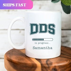 May include: White ceramic mug featuring the teal text "DDS", a progress bar, and the name "Samantha". The mug has a large handle and is on a wooden surface.