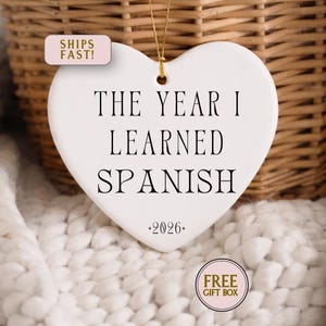 May include: A white heart-shaped ornament with the text "THE YEAR I LEARNED SPANISH" in black. The year "2026" is below the text. The ornament hangs from a gold string. A woven basket and a white, textured blanket are in the background.