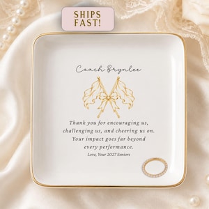 May include: A square white ceramic trinket dish with a gold-trimmed edge. The center features a gold graphic of two crossed flags tied with a bow, the text Coach Brynlee, and a message thanking a coach, signed Love, Your 2027 Seniors. A gold ring sits on the dish.