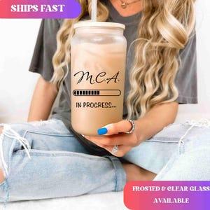 May include: A frosted glass tumbler with a bamboo lid and straw, filled with a creamy beverage. The tumbler features the text "M.C.A. IN PROGRESS..." in black script and a loading bar graphic. The glass is clear and available.