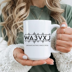 May include: White ceramic mug with a black graphic of a sound wave and the text "WA3VXJ BROADCASTING SINCE 2026". The mug has a white handle and is held by a person.