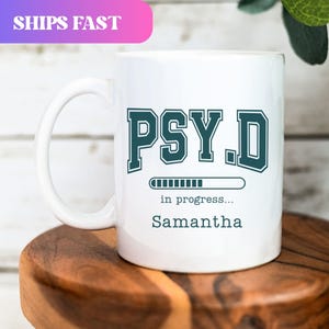 May include: White ceramic mug with a large handle. The mug features the text "PSY.D" in teal, with a progress bar and the name "Samantha" below. The top left corner has a pink and purple banner that says "SHIPS FAST".