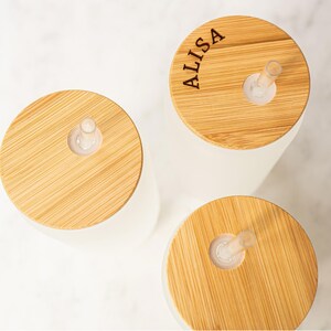 Puede incluir: Tres vasos de vidrio blancos con tapas de bamb&uacute; y pajitas transparentes. Una tapa est&aacute; grabada con el nombre "ALISA". Las tapas son de color marr&oacute;n claro y los vasos son blancos. Las pajitas son transparentes.