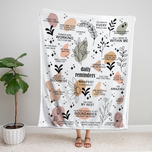 May include: White throw blanket with inspirational quotes and floral designs. The blanket features phrases like "I am amazing" and "Manifest it into reality." The design includes various colors and shapes.