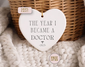 May include: A white heart-shaped ornament with the text "THE YEAR I BECAME A DOCTOR" in black. The year "2025" is printed below the text. A gold string is attached for hanging. The ornament is set against a soft, white background.