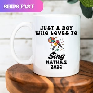 May include: White ceramic mug with the text "JUST A BOY WHO LOVES TO Sing NATHAN -2024-" and a graphic of microphones and musical notes. The mug also has a handle and is on a wooden surface.