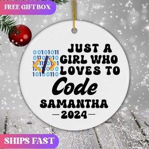 May include: A white round ornament with the text "JUST A GIRL WHO LOVES TO Code SAMANTHA -2024-". The ornament has a gold string for hanging and a binary code and coding symbols design. The ornament is decorated with a red ball and green leaves.