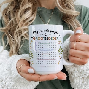 May include: White ceramic mug with the text "My favorite people call me Grootmoeder" and a word search puzzle with names. The mug has a large handle and is decorated with floral and heart accents.