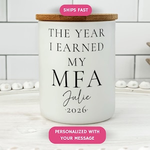 May include: A white ceramic jar with a bamboo lid. The jar has black text that reads "THE YEAR I EARNED MY MFA Julie 2026." The jar is on a white surface with decorative elements.