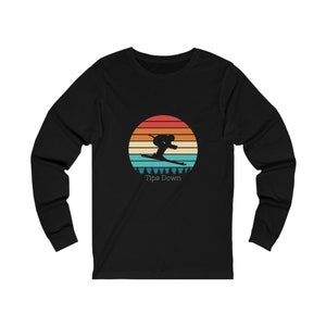 Op de afbeelding: Zwart shirt met lange mouwen met een retro ontwerp van een skiër tegen een zonsondergang. Het ontwerp bevat een cirkel met oranje, gele en blauwgroene strepen, met de tekst "Tips Down" eronder.