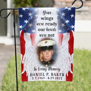 May include: A garden flag featuring a red, white and blue American flag design. A photograph of a man in military uniform is positioned in the centre of the flag. The text "Your wings were ready our hearts were not" is above the photograph. The text "In Loving Memory DANIEL P. BAKER 3.7.1969 - 8.29.2022" is below the photograph.