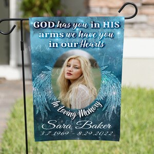 May include: A blue and white garden flag with a photo of a woman in a circle surrounded by angel wings. The text reads "GOD has you in HIS arms we have you in our Hearts In Loving Memory Sara Baker 3.7.1969 - 8.29.2022".