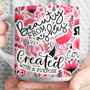 May include: White ceramic mug with a pink floral pattern. The mug has a black sticker with the text "Beauty from ashes" and a smaller sticker with the text "Isaiah 61:3". Another sticker says "Created with a purpose".