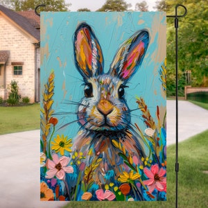 May include: A colorful painting of a rabbit with large ears surrounded by flowers. The rabbit is painted in shades of gray, brown, and blue, with bright yellow, pink, and blue flowers surrounding it. The background is a light blue.