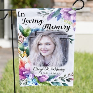 May include: A white garden flag with a watercolor floral design and a photo of a woman. The text "In Loving Memory" is at the top of the flag. The text "Cheryl R. Mackey 7.3.1942 - 4.30.23" is at the bottom of the flag.
