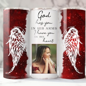 May include: A white tumbler with a red and white angel wing design. The tumbler has a photo of a woman and the text "God has you in his arms I have you in my heart".