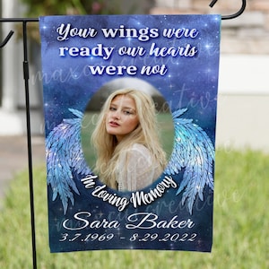 May include: A blue and white garden flag with a photo of a woman and angel wings. The text reads "Your wings were ready our hearts were not In Loving Memory Sara Baker 3.7.1969 - 8.29.2022".