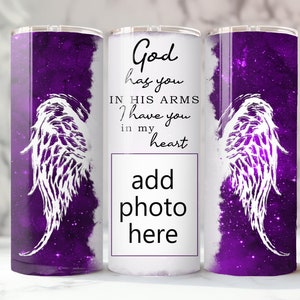 May include: A white tumbler with a purple galaxy background and white angel wings. The text "God has you in his arms I have you in my heart" is printed on the tumbler. There is a space to add a photo.