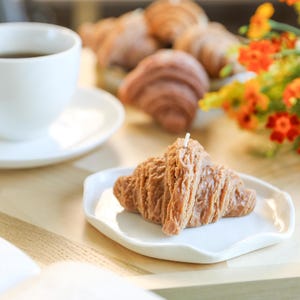 以下が含まれることがあります： 白い皿の上に置かれた、茶色のクロワッサン型のろうそく。ろうそくは、コーヒーカップとオレンジ色の花束が置かれた木製のテーブルの上にあります。