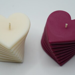 May include: Two heart-shaped candles, one white and one burgundy, are stacked in a spiral pattern. Each candle has a single wick.