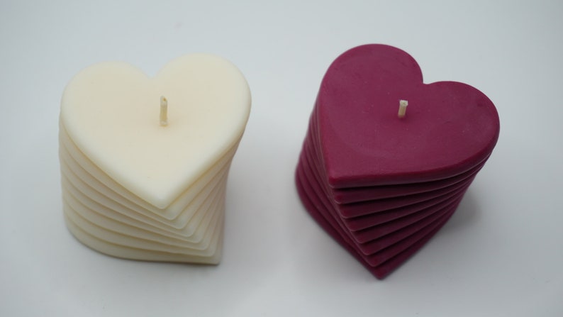 May include: Two heart-shaped candles, one white and one burgundy, are stacked in a spiral pattern. Each candle has a single wick.