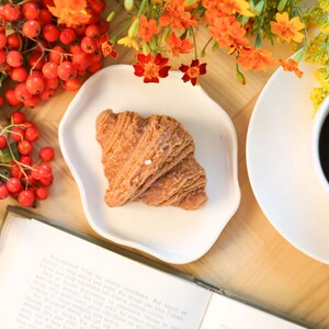 以下が含まれることがあります： 木製のテーブルの上に、クロワッサンが載った白い皿、コーヒーカップ、本が置かれています。背景には赤いベリーと黄色い花があります。