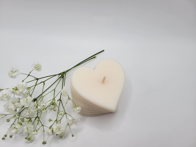May include: A white heart-shaped candle with a wick in the center, surrounded by a small bouquet of white baby's breath flowers.