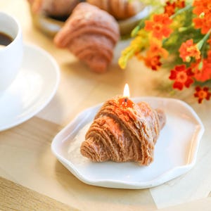 以下が含まれることがあります： 白い皿の上に置かれた、点火された芯を持つ茶色のクロワッサン型のろうそく。ろうそくは、コーヒーカップとオレンジ色の花束が背景にある木製の表面の上に置かれています。