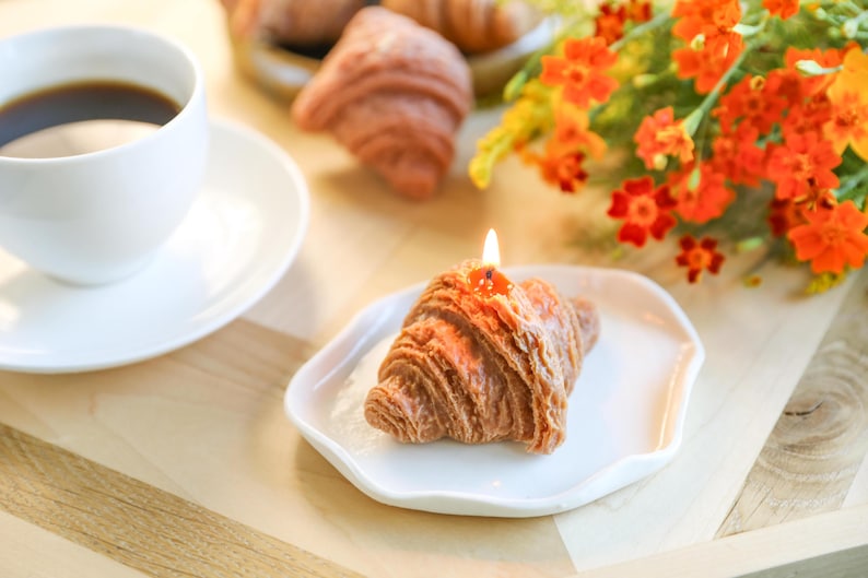 以下が含まれることがあります： 白い皿の上に置かれた、点火された芯を持つ茶色のクロワッサン型のろうそく。ろうそくは、コーヒーカップとオレンジ色の花束が背景にある木製の表面の上に置かれています。