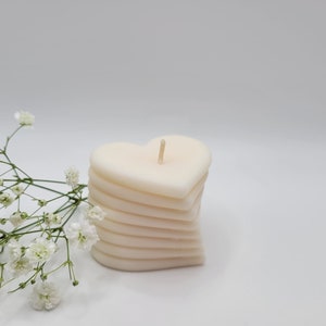 May include: A white heart-shaped candle with multiple layers. The candle is sitting on a white surface with a sprig of white flowers.